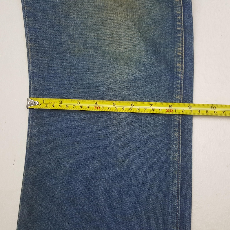 May include: Close-up of blue denim fabric with a yellow measuring tape. The tape shows measurements in centimetres, highlighting the width of the denim. The fabric has a faded appearance, with visible seams.