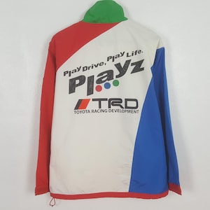 Puede incluir: Chaqueta vintage con dise&ntilde;o de bloques de color en rojo, blanco y azul. La parte trasera presenta el texto "Play Drive, Play Life" y "Playz" con el logo TRD Toyota Racing Development. El cuello es verde.