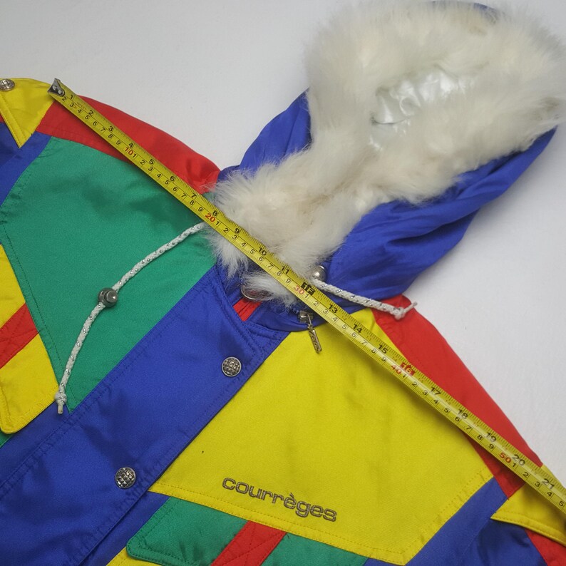 May include: A colorful vintage winter jacket with a fur-lined hood. The jacket features a patchwork design in red, yellow, green, and blue. The brand name "courr&egrave;ges" is visible on the front. A measuring tape is laid across the jacket.