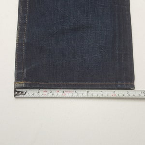 May include: Close-up of dark blue denim jeans with a measuring tape showing a width of approximately 25 cm. The jeans have visible stitching and a slightly worn appearance, suggesting a vintage or well-loved item.