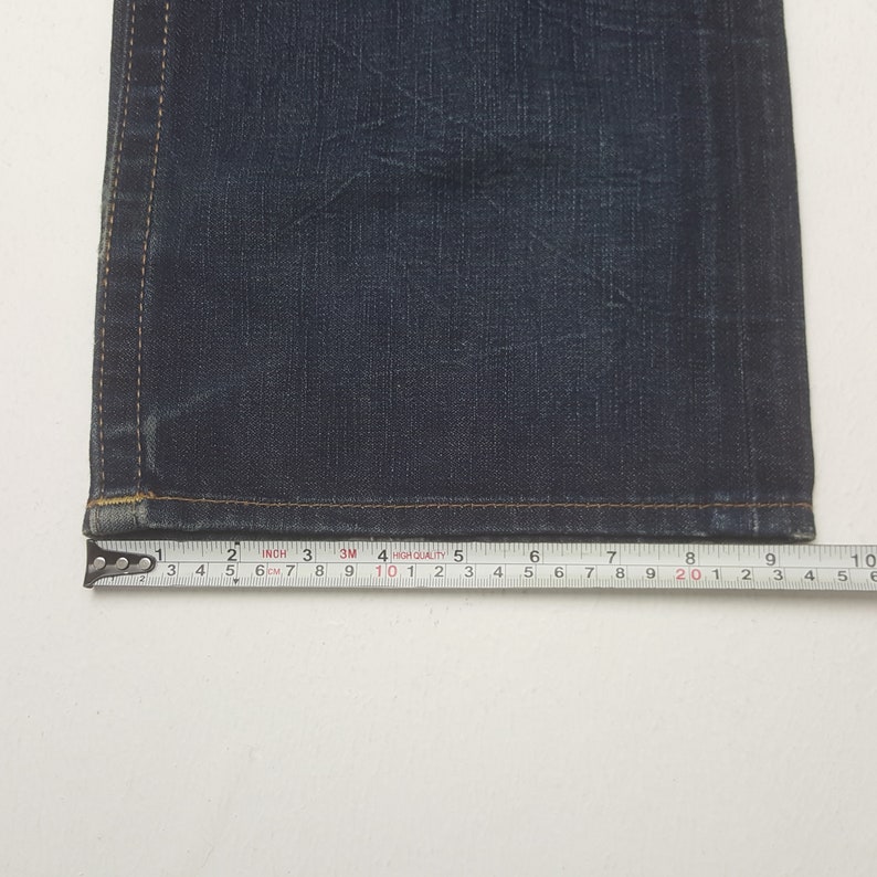 May include: Close-up of dark blue denim jeans with a measuring tape showing a width of approximately 25 cm. The jeans have visible stitching and a slightly worn appearance, suggesting a vintage or well-loved item.