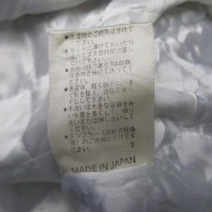 May include: A close-up of a white fabric tag with Japanese text and the words "MADE IN JAPAN" in black. The tag provides care instructions, including washing and drying advice. The fabric appears to be quilted.