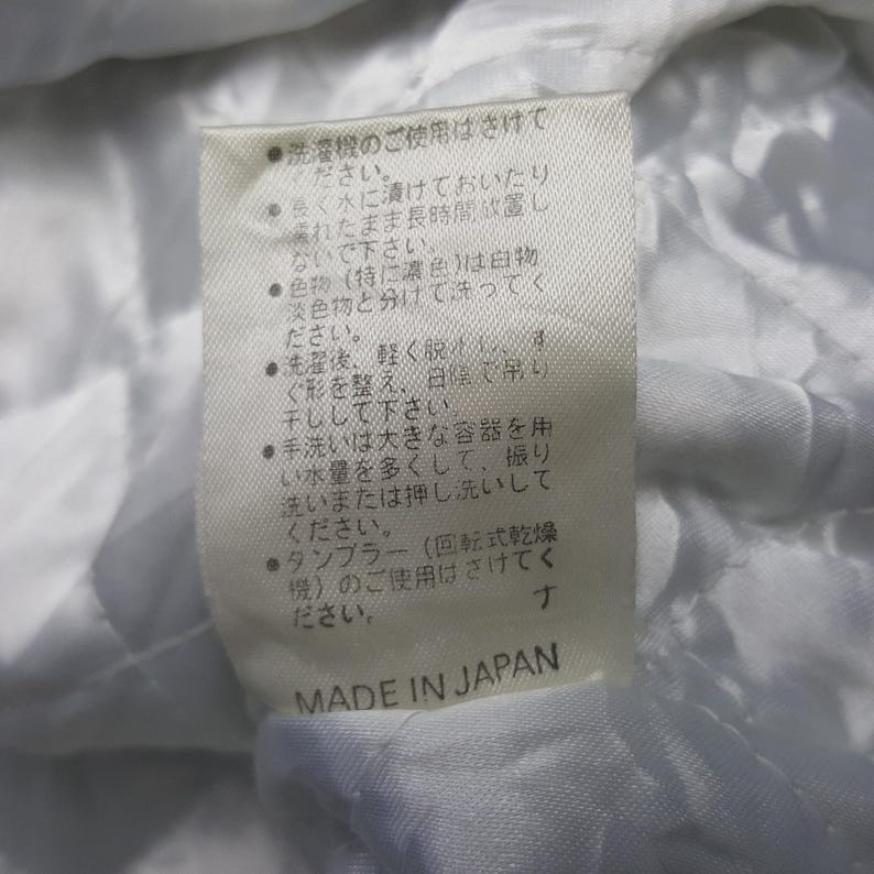 May include: A close-up of a white fabric tag with Japanese text and the words "MADE IN JAPAN" in black. The tag provides care instructions, including washing and drying advice. The fabric appears to be quilted.