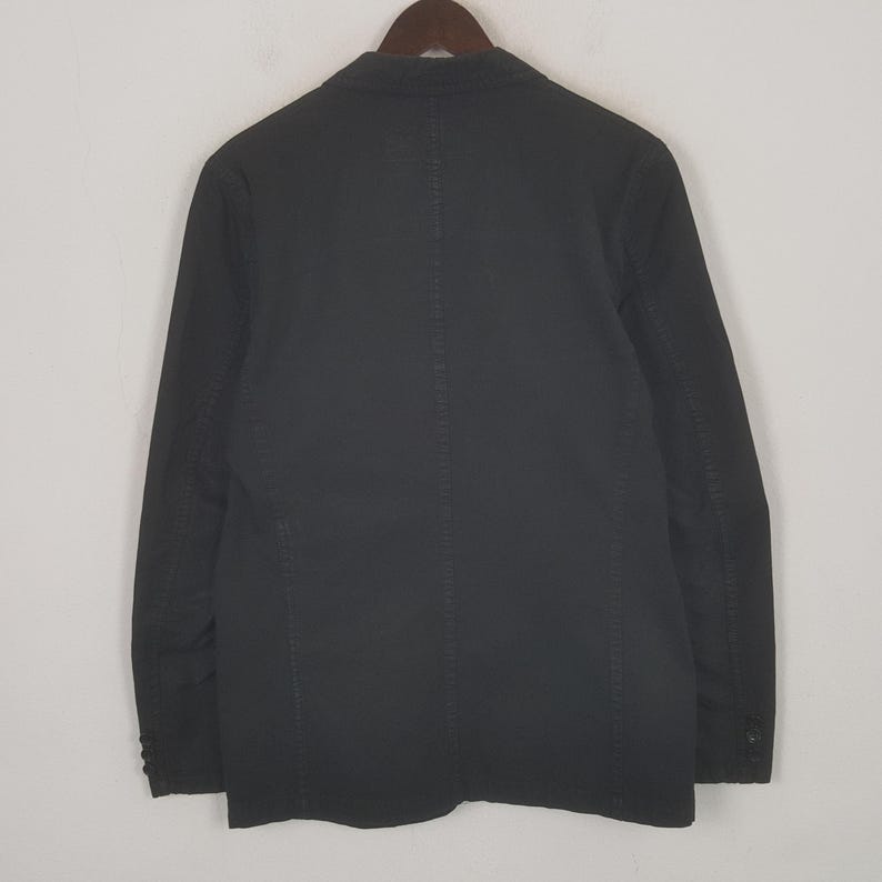 Puede incluir: Un blazer negro con cierre de dos botones y solapa con muescas. La chaqueta est&aacute; hecha de una tela oscura y lisa y tiene un dise&ntilde;o cl&aacute;sico y entallado. El blazer est&aacute; colgado de una percha de madera.