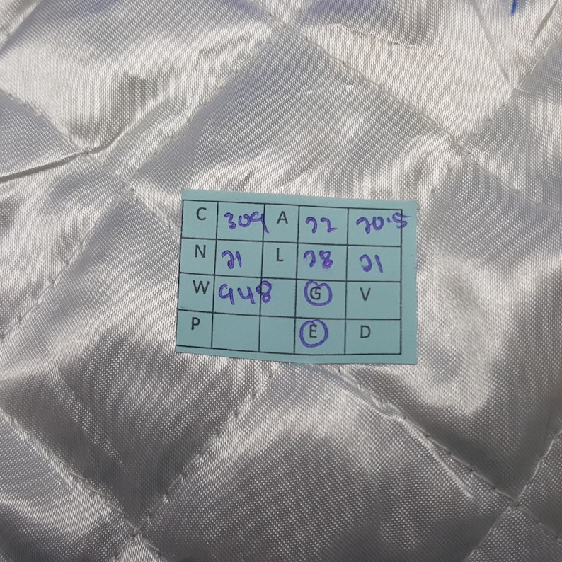 May include: A close-up of a quilted, off-white fabric. A small, light blue tag with handwritten numbers and letters is attached to the fabric. The fabric has a diamond pattern.