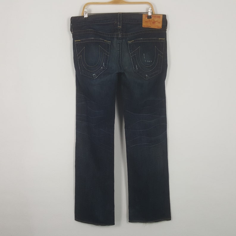 May include: Dark wash denim jeans with a straight leg cut, hanging on a wooden hanger. The jeans feature distressed details and a leather patch on the back. The back pockets have a distinctive stitching design.