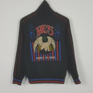 Könnte beinhalten: Schwarze Jacke mit Reißverschluss und roten und blauen Streifen an den Bündchen und Ärmeln. Auf dem Rücken befindet sich eine Grafik mit dem Text "MC5" über einem Adlerdesign und den Worten "BABES IN ARMS".