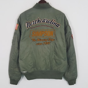 May include: Olive green bomber jacket with embroidered text on the back reading "North Carolina" and "Simpson". The jacket has a ribbed collar, cuffs, and hem, with a small American flag patch on the left sleeve.