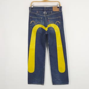 Puede incluir: Vaqueros de mezclilla azul oscuro con un gran gráfico amarillo en la parte trasera que se asemeja a los arcos dorados de McDonald's. Los vaqueros cuelgan de una percha de madera, mostrando los bolsillos traseros y una etiqueta roja de Levi's.
