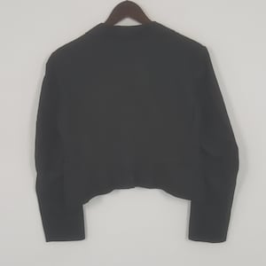 May include: A black blazer hanging on a wooden hanger. The blazer is a solid color with long sleeves and a tailored fit. The back of the blazer is visible, showing the cut and design.