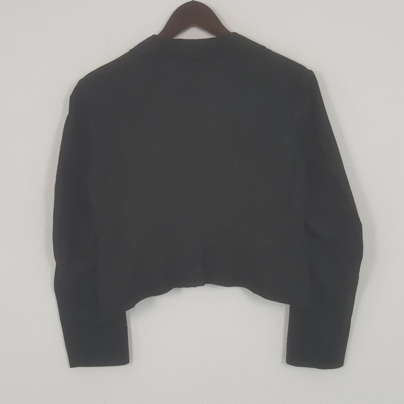 May include: A black blazer hanging on a wooden hanger. The blazer is a solid color with long sleeves and a tailored fit. The back of the blazer is visible, showing the cut and design.