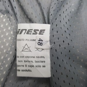 Peut inclure: Gros plan sur une &eacute;tiquette de v&ecirc;tement avec le nom de marque "INESE" en blanc. L'&eacute;tiquette indique les instructions de lavage et la taille "48". Le tissu est un mat&eacute;riau en maille gris clair.