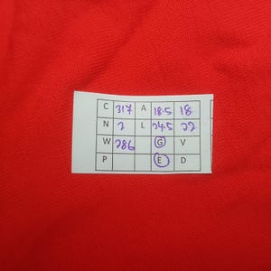 May include: A close-up of a red fabric background with a white tag. The tag has handwritten text and numbers in black ink. The tag includes the following text: C 317, A 18.5, 18, N 2, L 24.5, 22, W 286, G, E, D.