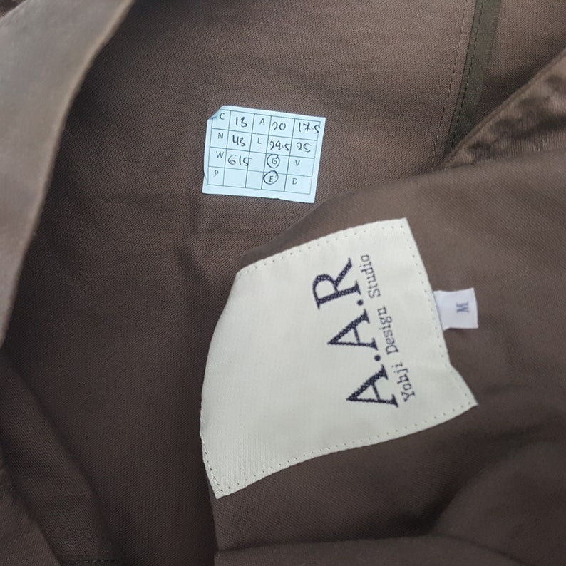 Puede incluir: Primer plano de una prenda marr&oacute;n con una etiqueta blanca que dice "A.A.R Yohji Design Studio". Una etiqueta blanca m&aacute;s peque&ntilde;a con la letra "M" es visible. Tambi&eacute;n es visible una peque&ntilde;a etiqueta blanca con medidas.