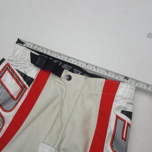 Peut inclure: Gros plan sur un pantalon de moto blanc et rouge avec des accents noirs. Le pantalon a une ceinture noire et une fermeture boutonn&eacute;e. Un m&egrave;tre ruban indique que la taille mesure environ 33 cm.