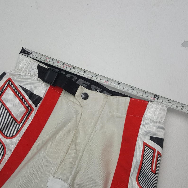 Peut inclure: Gros plan sur un pantalon de moto blanc et rouge avec des accents noirs. Le pantalon a une ceinture noire et une fermeture boutonn&eacute;e. Un m&egrave;tre ruban indique que la taille mesure environ 33 cm.