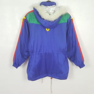 May include: A vintage blue ski jacket with a hood trimmed with white faux fur. The jacket features red and yellow accents on the sleeves and shoulders, with a green panel across the upper back. A drawstring cinches the waist. A retro winter coat.