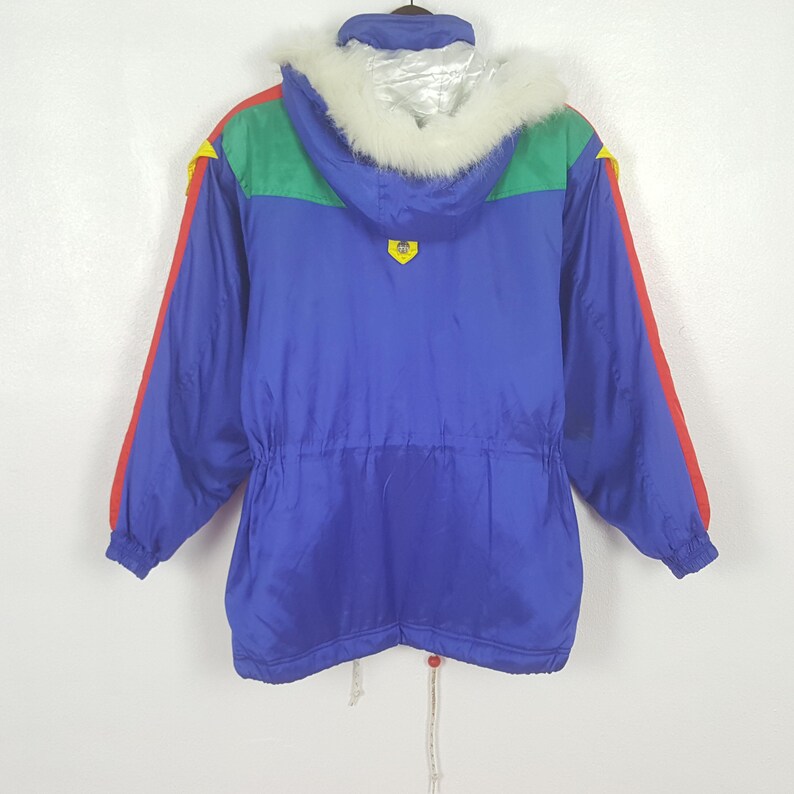 May include: A vintage blue ski jacket with a hood trimmed with white faux fur. The jacket features red and yellow accents on the sleeves and shoulders, with a green panel across the upper back. A drawstring cinches the waist. A retro winter coat.