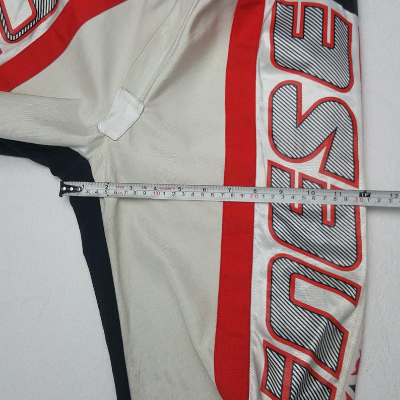 Peut inclure: Gros plan sur une jambe de pantalon de course blanche et rouge avec le mot "DUSE" en grandes lettres ray&eacute;es noires et blanches. Une bande noire longe le c&ocirc;t&eacute;. La mesure est d'environ 33 cm.