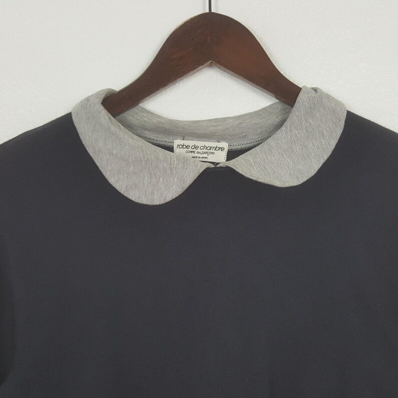 K&ouml;nnte beinhalten: Ein dunkelgraues T-Shirt mit einem hellgrauen Bubikragen. Der Kragen hat eine Kontrastfarbe zum Shirt. Das Etikett lautet "robe de chambre COMME des GAR&Ccedil;ONS". Das Shirt h&auml;ngt an einem Holzkleiderb&uuml;gel.