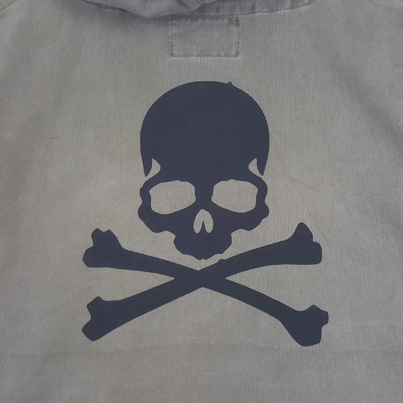 Puede incluir: Una sudadera con capucha gris con un dise&ntilde;o de calavera y huesos cruzados azul oscuro en la espalda. La calavera tiene cuencas oculares grandes y una nariz prominente. Los huesos cruzados est&aacute;n colocados debajo de la calavera, creando un dise&ntilde;o cl&aacute;sico de tem&aacute;tica pirata.
