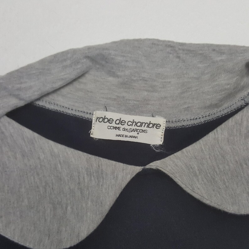 K&ouml;nnte beinhalten: Nahaufnahme eines marineblauen und grauen Hemdes mit Kragen. Der Kragen ist grau, darunter ein marineblaues Band. Ein Etikett lautet "robe de chambre COMME des GARCONS MADE IN JAPAN."