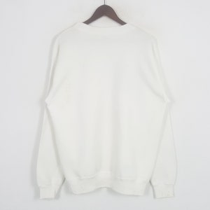 May include: A white crew neck sweatshirt hanging on a wooden hanger. The sweatshirt has long sleeves and ribbed cuffs and hem. The fabric appears to be a medium weight, suitable for cooler weather.