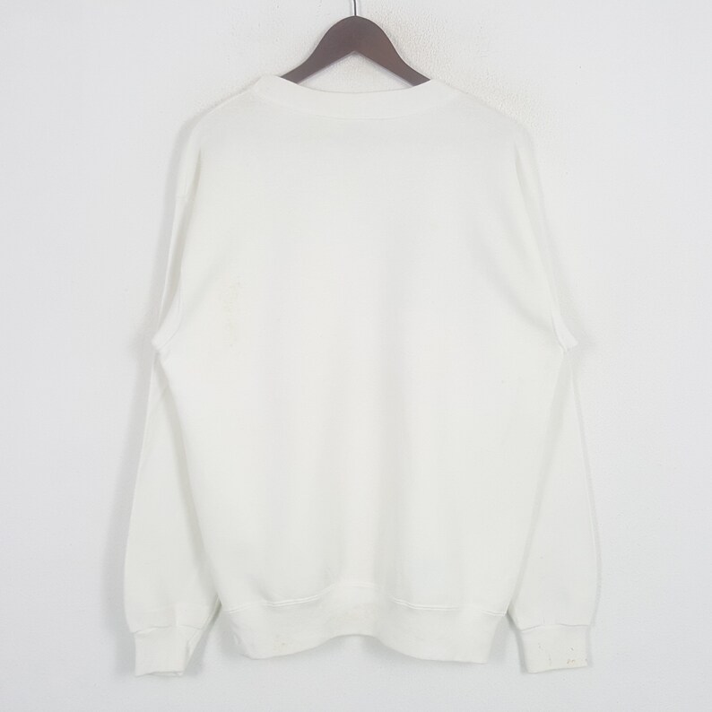 May include: A white crew neck sweatshirt hanging on a wooden hanger. The sweatshirt has long sleeves and ribbed cuffs and hem. The fabric appears to be a medium weight, suitable for cooler weather.