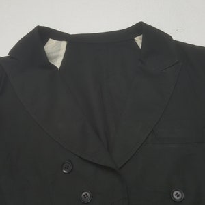 May include: A close-up of a black blazer with a double-breasted design. The blazer features a notched lapel, two rows of buttons, and a tailored fit. The fabric appears to be a smooth, solid color, and the garment is likely made of a lightweight material.