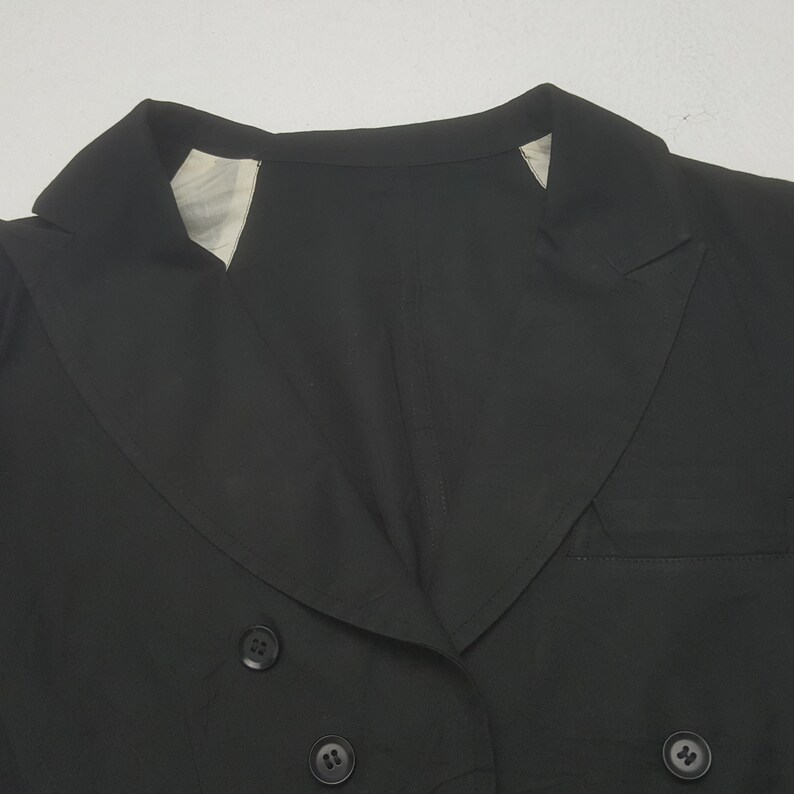 May include: A close-up of a black blazer with a double-breasted design. The blazer features a notched lapel, two rows of buttons, and a tailored fit. The fabric appears to be a smooth, solid color, and the garment is likely made of a lightweight material.