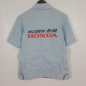 May include: Light blue short-sleeved shirt with the text "MUGEN 無限 HONDA" printed on the back in black and red. The shirt has a collar and red trim along the bottom hem and sleeve cuffs. The shirt is hanging on a wooden hanger.