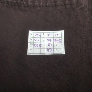 Puede incluir: Primer plano de una tela marr&oacute;n oscuro con una etiqueta rectangular blanca. La etiqueta contiene n&uacute;meros y letras manuscritos en tinta morada, incluyendo "294", "21", "18", "32", "29" y "25.5". La tela es de color liso.