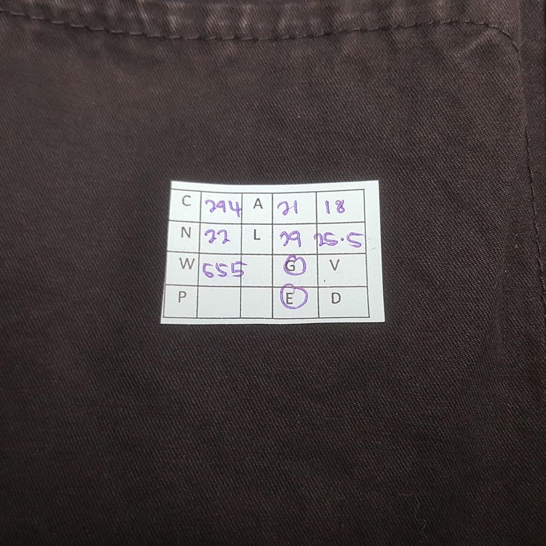 Puede incluir: Primer plano de una tela marr&oacute;n oscuro con una etiqueta rectangular blanca. La etiqueta contiene n&uacute;meros y letras manuscritos en tinta morada, incluyendo "294", "21", "18", "32", "29" y "25.5". La tela es de color liso.