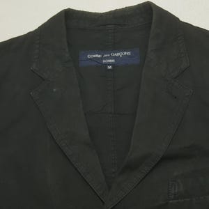 Puede incluir: Blazer negro con solapas de muesca y cierre de un solo bot&oacute;n. El blazer presenta una etiqueta con el texto "COMME des GAR&Ccedil;ONS HOMME" y la talla "M". La chaqueta est&aacute; hecha de una tela oscura lisa.