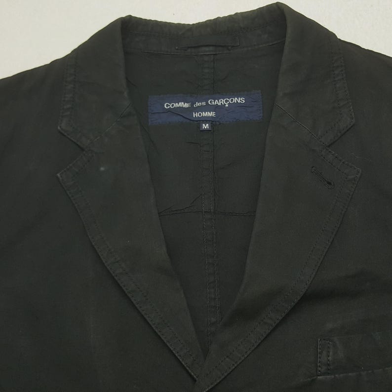 Puede incluir: Blazer negro con solapas de muesca y cierre de un solo bot&oacute;n. El blazer presenta una etiqueta con el texto "COMME des GAR&Ccedil;ONS HOMME" y la talla "M". La chaqueta est&aacute; hecha de una tela oscura lisa.