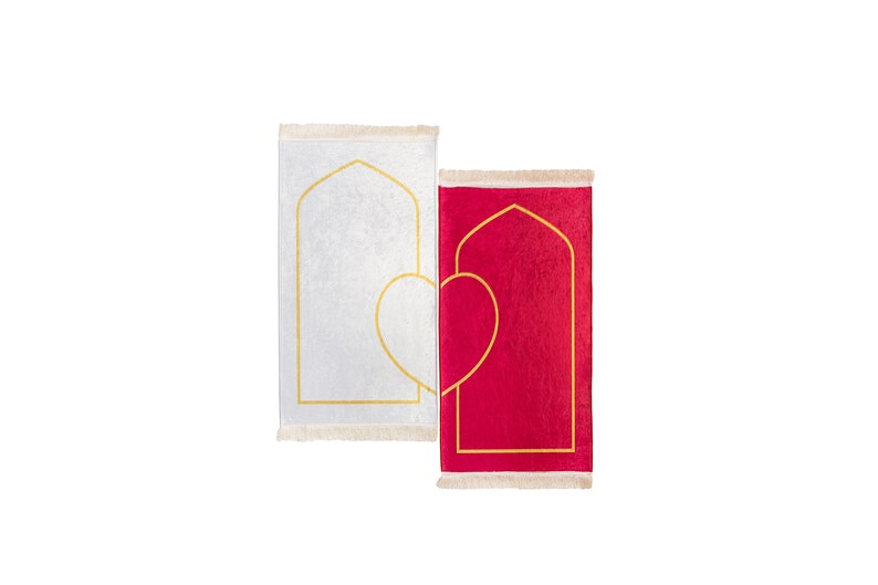 May include: Two prayer rugs, one white and one red, are displayed side by side. Each rug features a gold arched design and a gold heart shape. The rugs have a fringed edge.