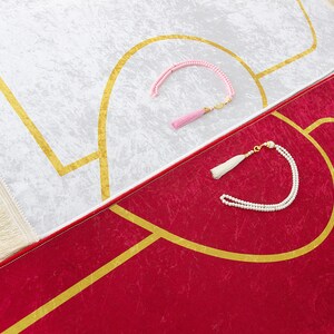 May include: Two prayer rugs, one white and one red, with gold geometric designs. The white rug has a pink tassel prayer bead, and the red rug has a white tassel prayer bead. Both rugs have a fringed border.