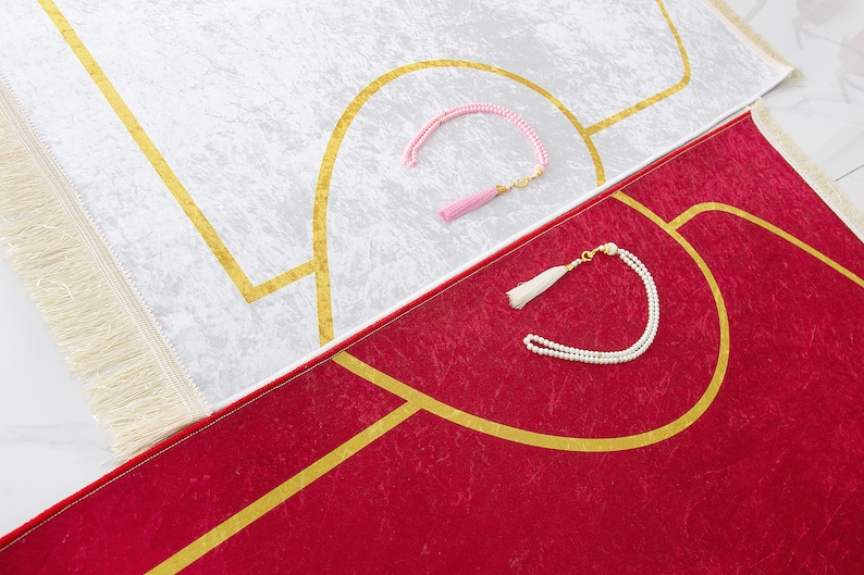 May include: Two prayer rugs, one white and one red, with gold geometric designs. The white rug has a pink tassel prayer bead, and the red rug has a white tassel prayer bead. Both rugs have a fringed border.