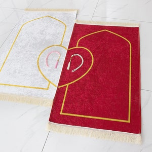 May include: Two prayer rugs, one white and one red, are displayed. Each rug features a gold-colored arched design and a heart-shaped accent. The rugs have a fringed border and include a small tassel. The rugs are placed on a white tiled surface.