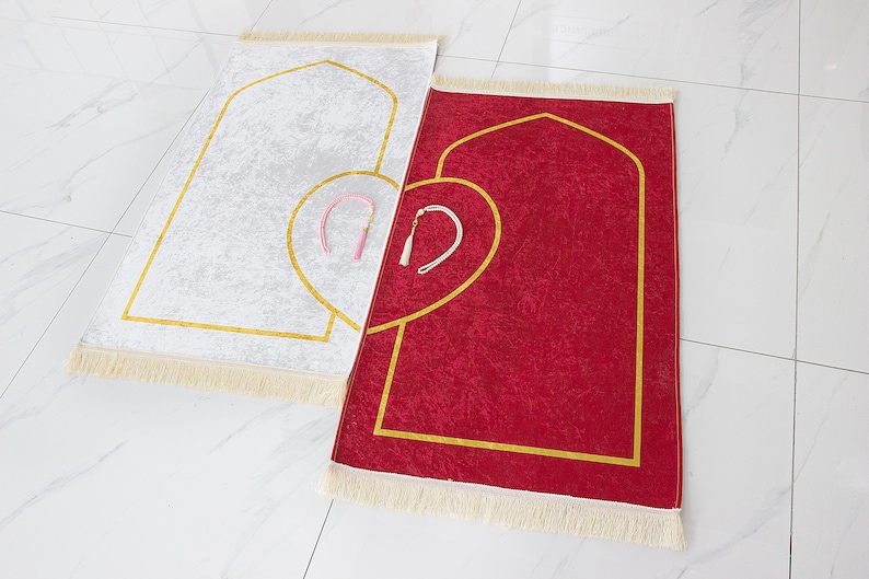 May include: Two prayer rugs, one white and one red, are displayed. Each rug features a gold-colored arched design and a heart-shaped accent. The rugs have a fringed border and include a small tassel. The rugs are placed on a white tiled surface.