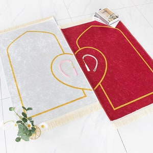 May include: Two prayer rugs, one silver and one red, are displayed. Both have a gold border and a heart-shaped design. Each rug has a tassel and a matching pink and white accessory. A small floral arrangement is in the lower left corner.