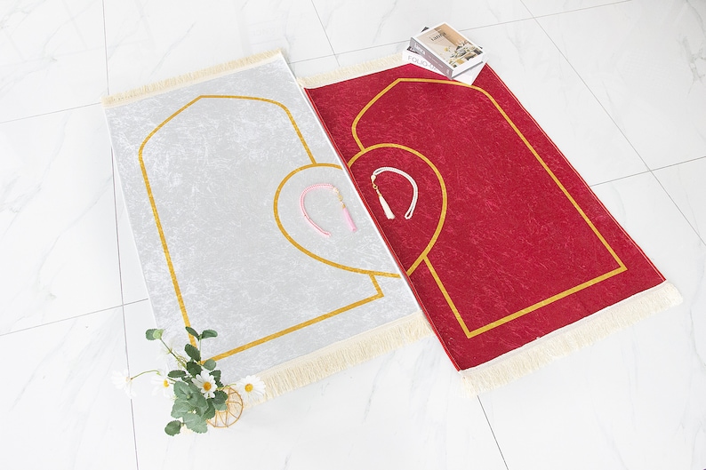 May include: Two prayer rugs, one silver and one red, are displayed. Both have a gold border and a heart-shaped design. Each rug has a tassel and a matching pink and white accessory. A small floral arrangement is in the lower left corner.
