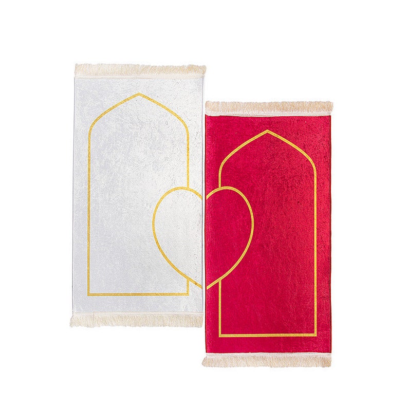May include: Two prayer rugs, one white and one red, are displayed side-by-side. Each rug features a gold-colored arched design with a heart shape. The rugs have a fringed edge. The white rug has a textured appearance.