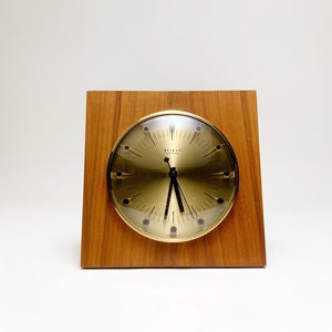 May include: A vintage desk clock with a gold-tone face and black hands, set within a square wooden frame. The clock face features a minimalist design with hour markers. The clock is a classic piece of home decor.