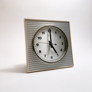 May include: A silver and white analog clock with black hands and black numbers on a white face. The clock is in a square, cream-colored frame with a striped pattern.