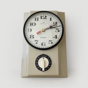 Puede incluir: Un reloj de pared de cuarzo beige con bisel negro y esfera blanca. El reloj tiene números y manecillas negros, con un segundero rojo. Un dial de temporizador está debajo de la esfera del reloj. El reloj mide aproximadamente 20 cm de alto.