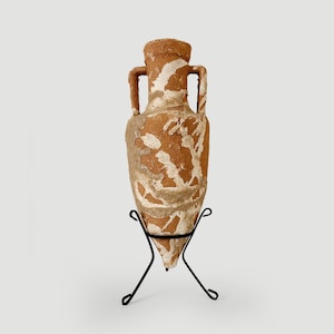 Può includere: Un'anfora in terracotta con una superficie ruvida e usurata caratterizzata da depositi minerali bianchi. Il vaso presenta due manici laterali e una base appuntita, sostenuto da un supporto in filo metallico nero in posizione verticale.