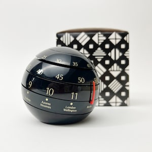 Könnte beinhalten: Ein schwarzer, kugelförmiger Timer mit goldenen Ziffern und einem roten Indikator. Der Timer zeigt Zeitzonen wie London, Wellington und Berlin an. Eine schwarz-weiß gemusterte Box befindet sich im Hintergrund.