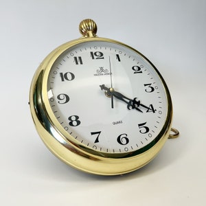 Puede incluir: Un reloj analógico dorado con agujas negras y números romanos. La esfera del reloj dice "Meister-Ankir" y "Quartz".