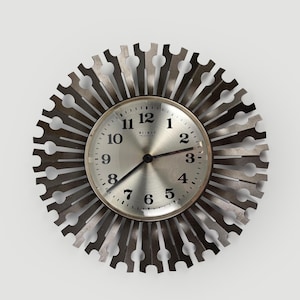 May include: A round wall clock with a metallic silver face featuring black Arabic numerals from 1 to 12 and the text WEIMAR. The clock is surrounded by a dark, sunburst-style frame made of radiating metal spokes with circular cutouts.